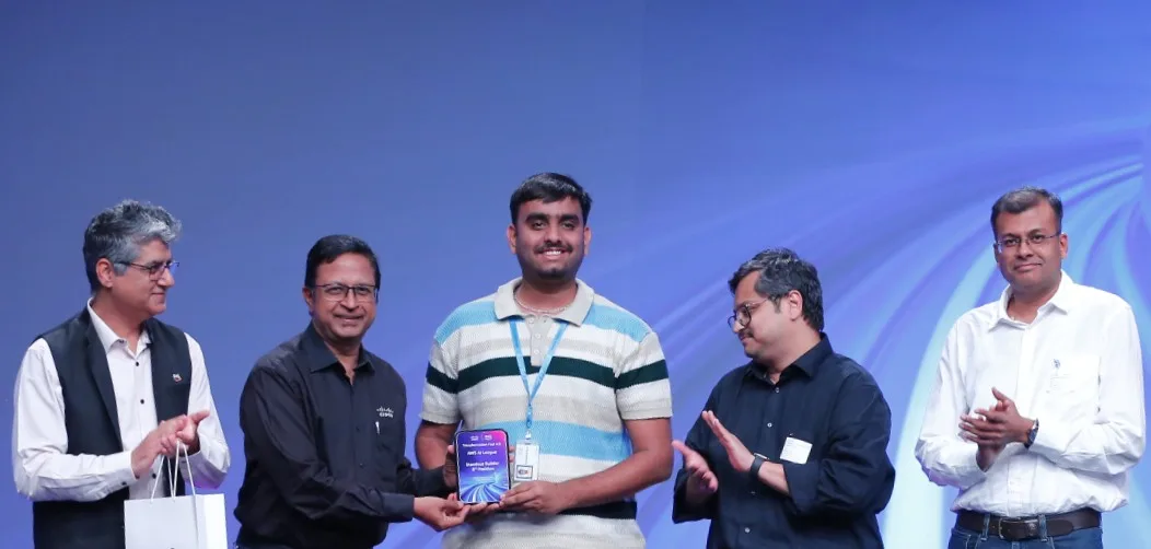On stage holding the award plaque at the Cisco × AWS AI hackathon, flanked by four Cisco leaders applauding.